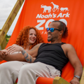 A couple laughing whilst sitting on an orange giant deck chair with a Noah's Ark Zoo Farm Logo on it. 