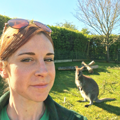 Female keeper wearing sunglasses on her head smiling at the camera with a wallaby standing on the grass looking at the camera.  
