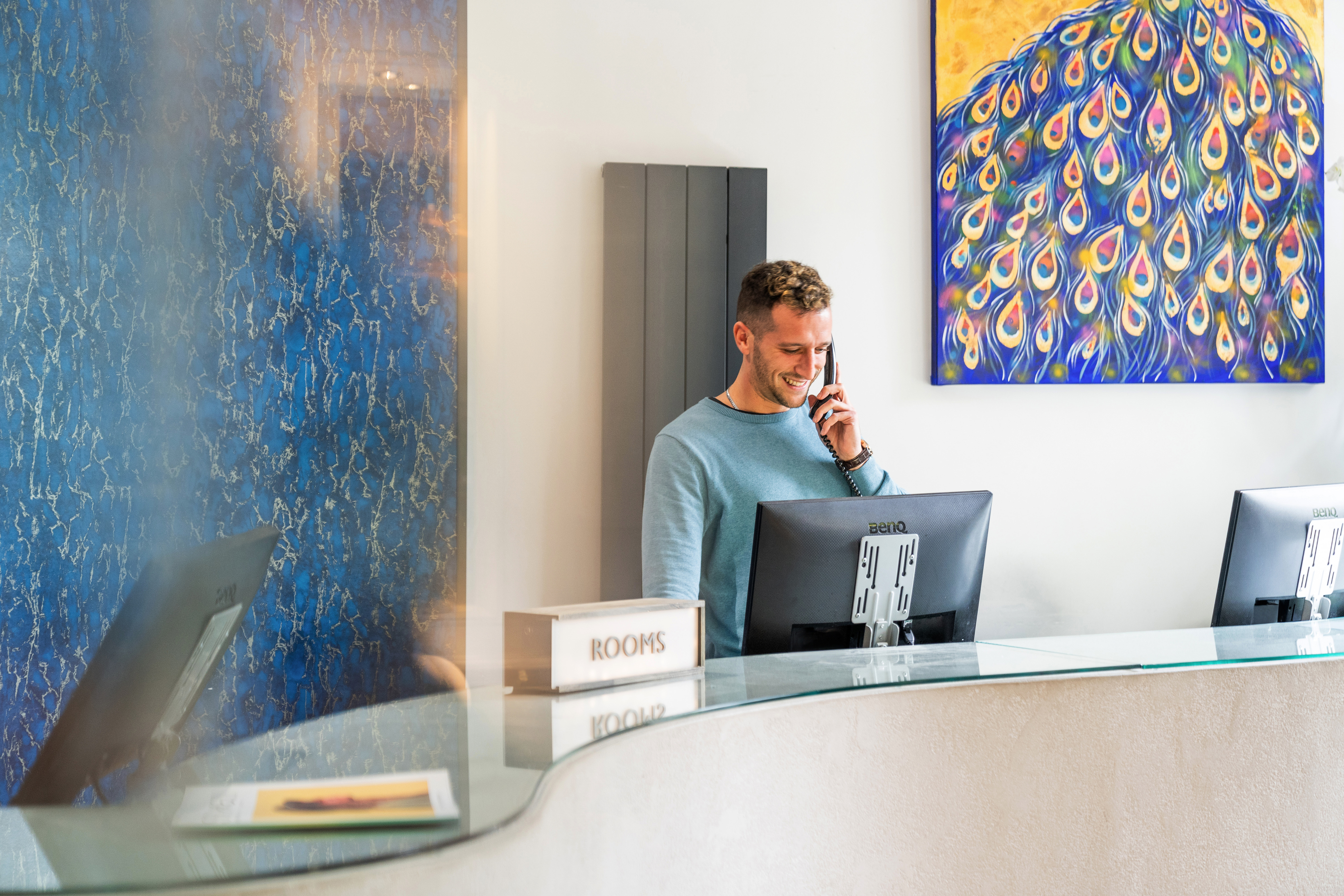 A smiling man is stood behind a reception desk on the phone. 
