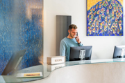 A smiling man is stood behind a reception desk on the phone. 
