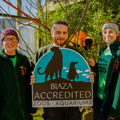 a male keeper holds a sign that says 'BIAZA Accredited', with two female keepers either side of him and a red ruffed lemur in the background.