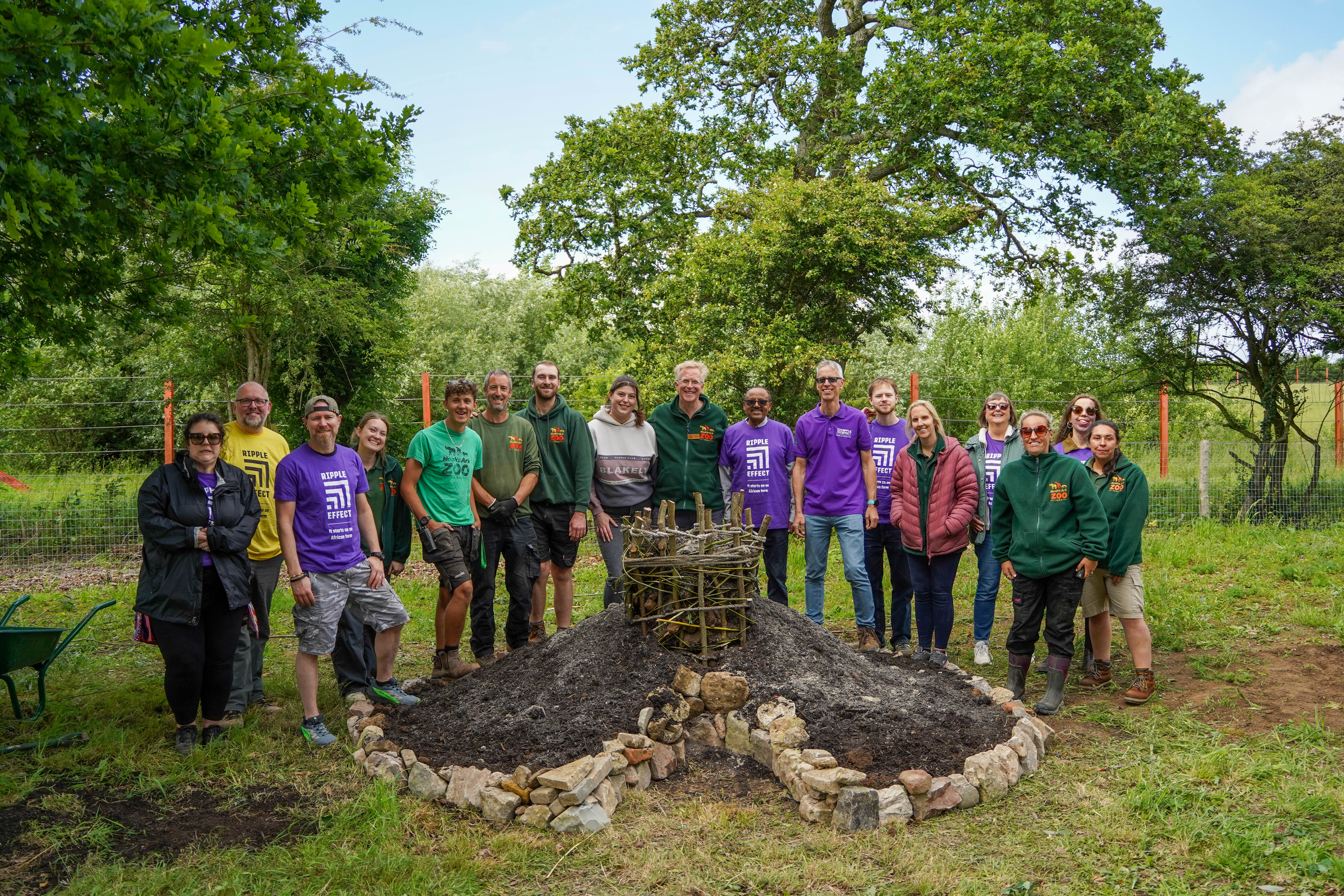 Team of people including staff in Noah's Ark uniform and Ripple effect Purple uniform stand behind a keyhole garden under a tree