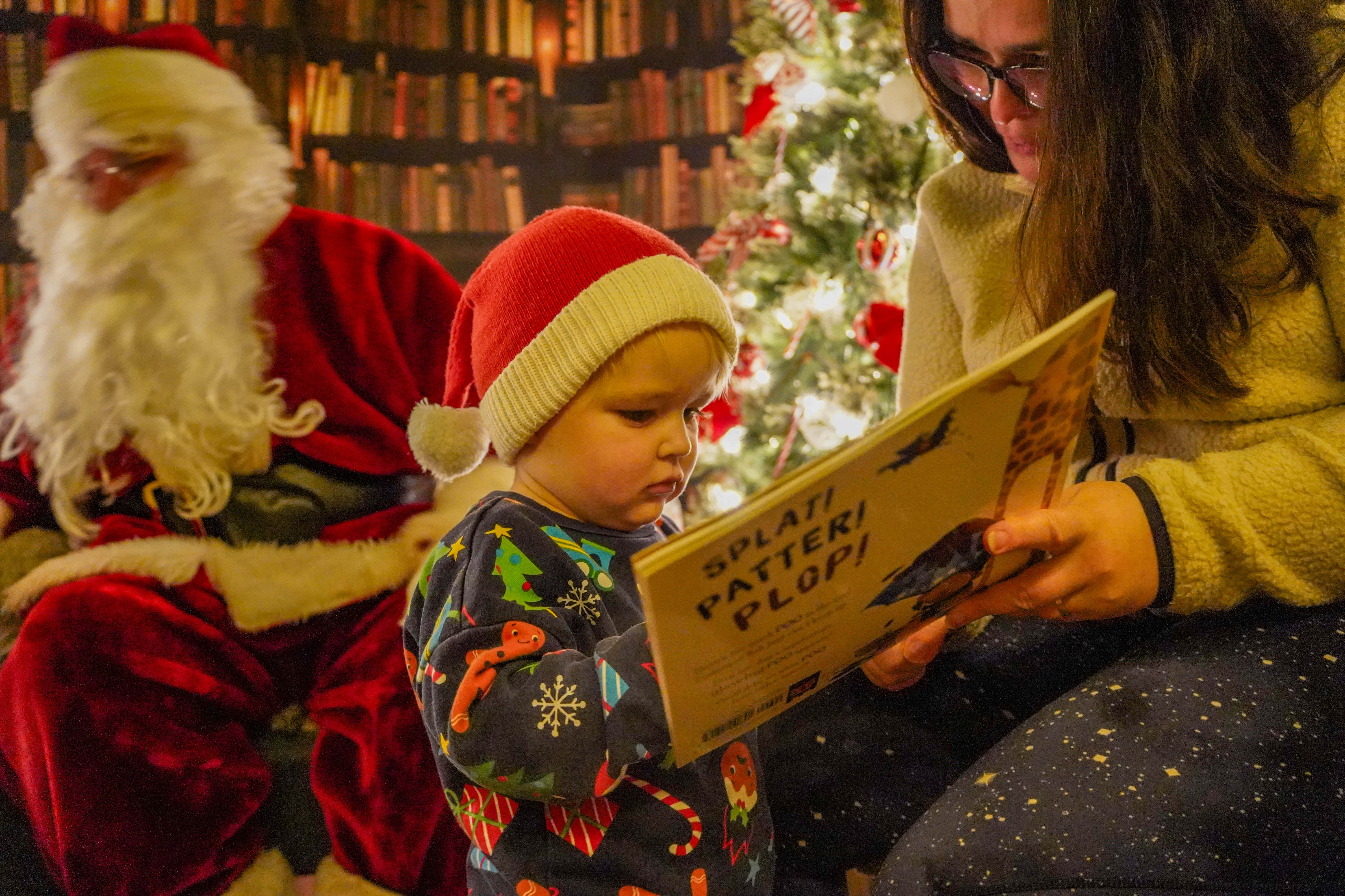 A little boy reading a book with his mum in Santa's library, with Santa behind them.