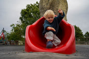 Toddler climbing out the bottom of a red slide with play park in background. 