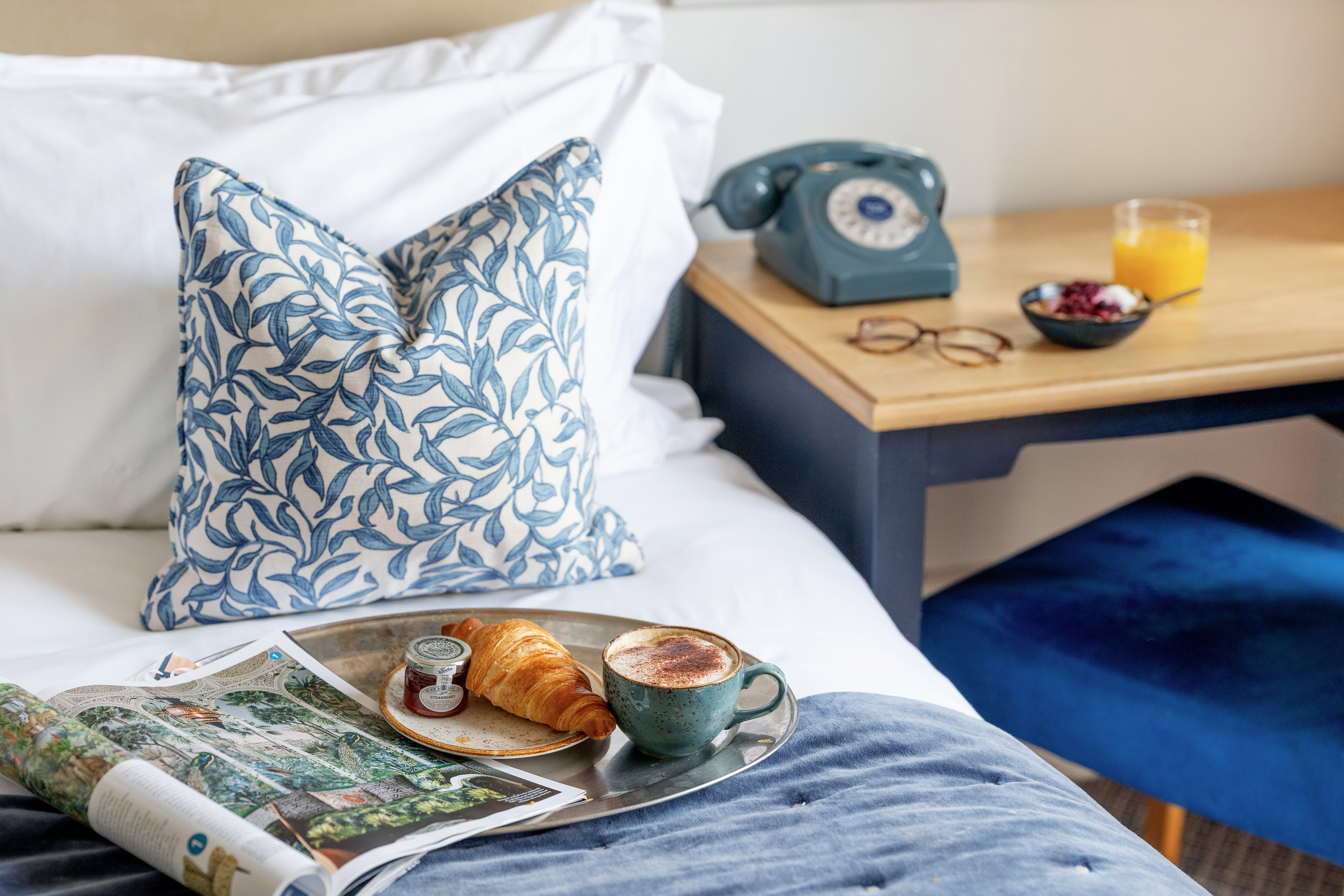 A croissant and a coffee, next to a magazine arranged on a bed with a blue floral cushion. The bed is next to a desk with a traditional telephone. 