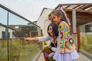 A young girl wearing a rainbow crochet cardigan and a purple skirt stands with her mother, pointing towards the glass window of the meerkat habitat.