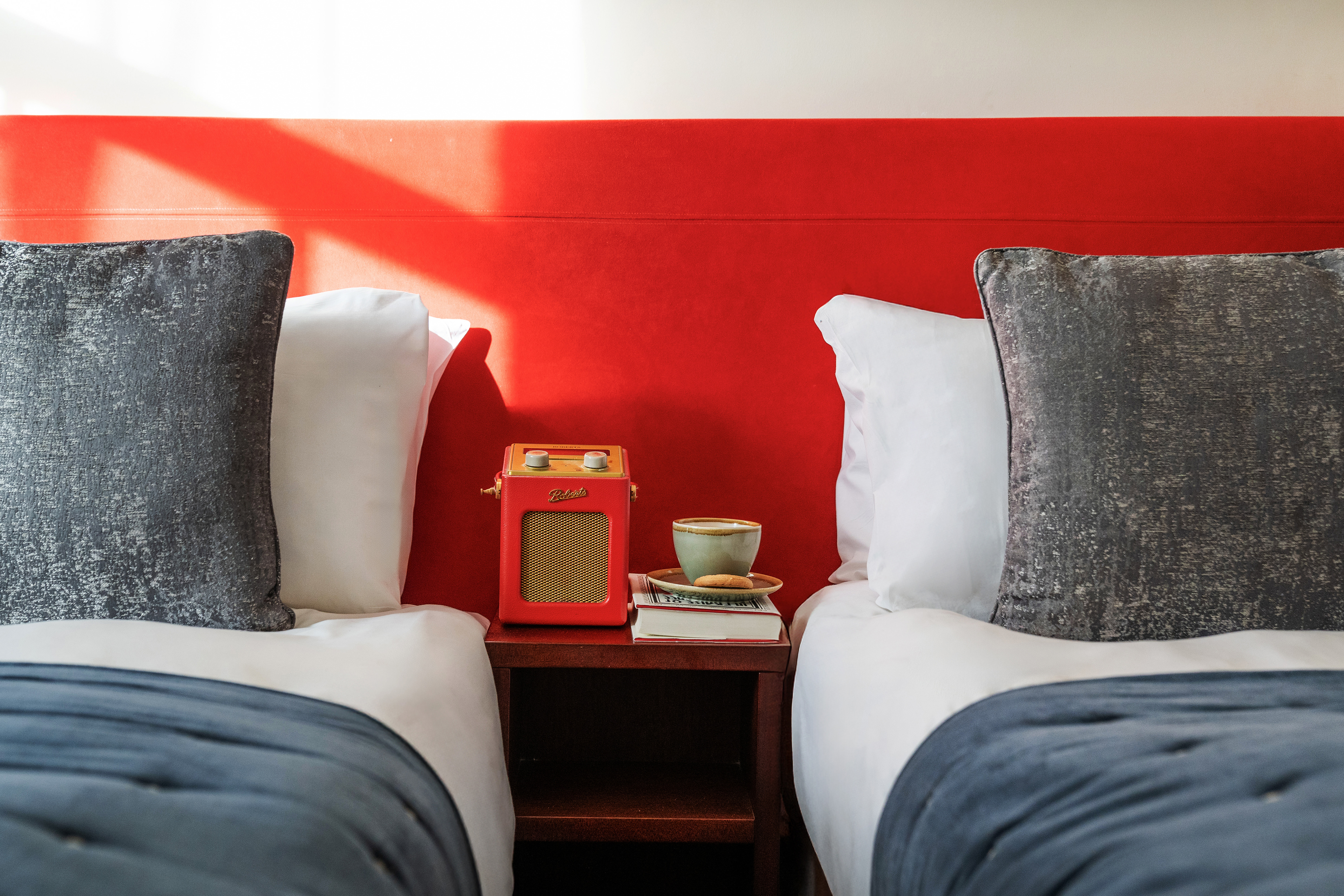 Luxurious twin beds against an orange wall with a radio and cup of tea and a book on a table between the beds. 