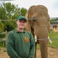 Keeper wearing a zoo branded jumper and cap, smiling at the camera, with a African Elephant behind him. 